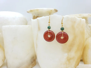 Image of gold pendant earrings with green bead and red bi disc hanging on white crystal.