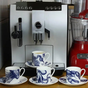 Set of four white espresso cups with blue nautical designs on saucers, displayed in front of espresso machine.