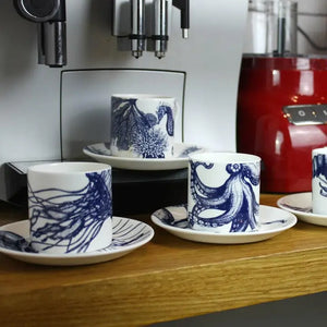 Set of four white espresso cups and saucers with blue marine life designs, displayed on wooden counter near espresso machine.