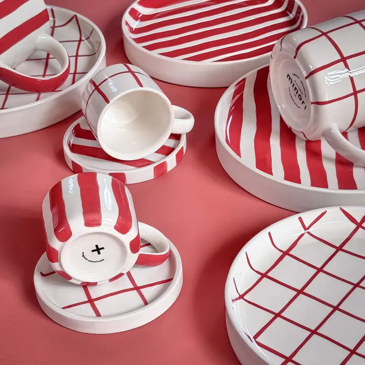 Red and white striped and checkered ceramic mugs and plates, arranged on a pink background.