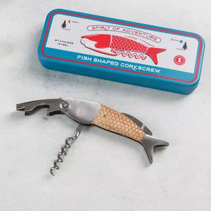 A fish-shaped corkscrew with a stainless steel body and a textured, scale-like handle is placed on a light surface. Above it is a rectangular blue tin box with red and white labeling that reads "SPIRIT OF ADVENTURE" at the top, featuring an image of a red fish. The text on the box also includes "STAINLESS STEEL," "FISH SHAPED CORKSCREW," and other small details.