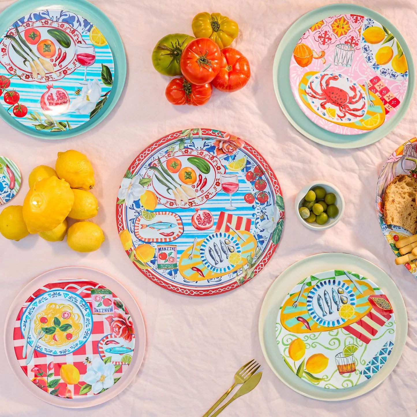 Colourful plates with food illustrations, surrounded by tomatoes, lemons, olives, breadsticks, and a fork on a tablecloth.