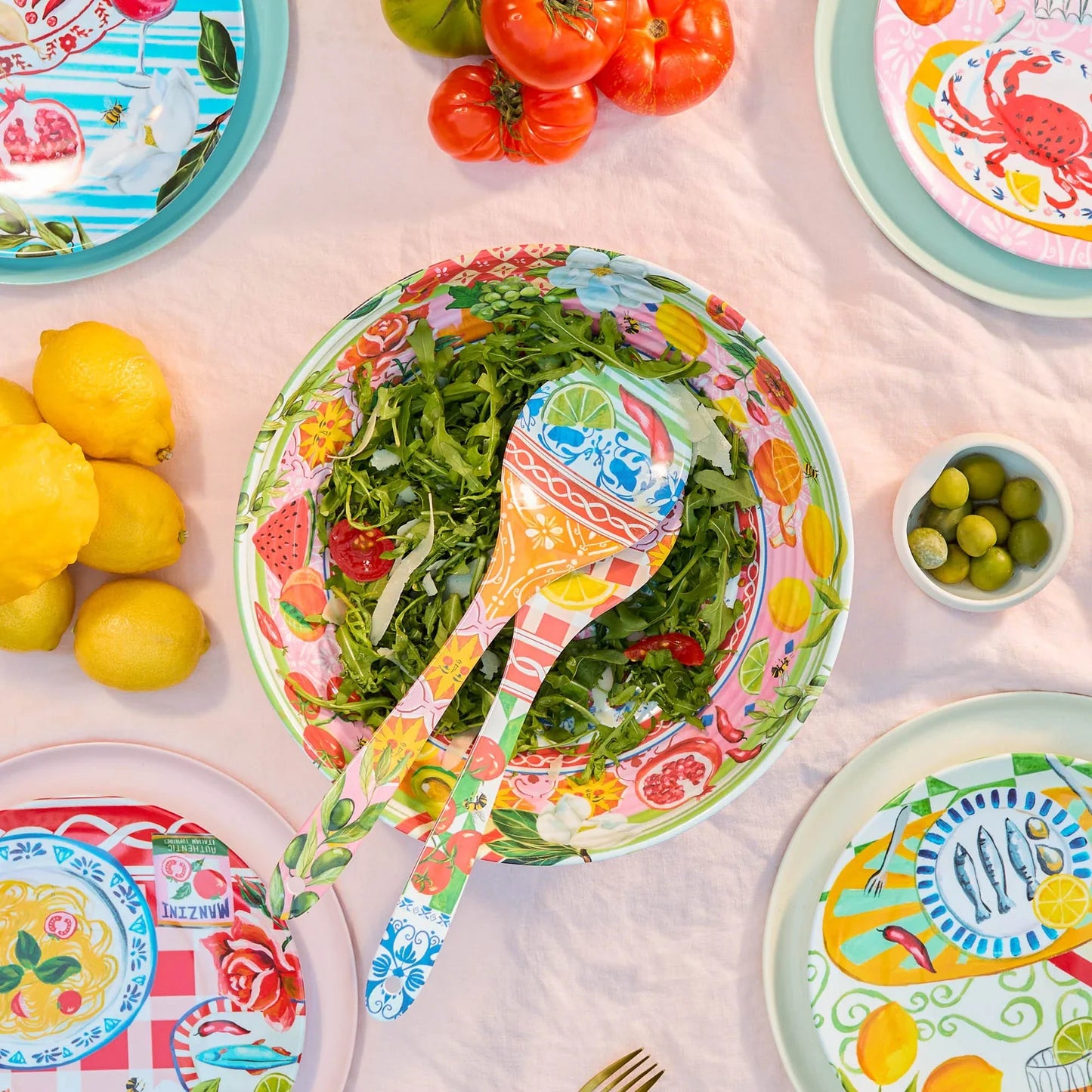 Italian summer table setting with salad bowl, utensils, lemons, tomatoes, olives, and vibrant plates on pink cloth.