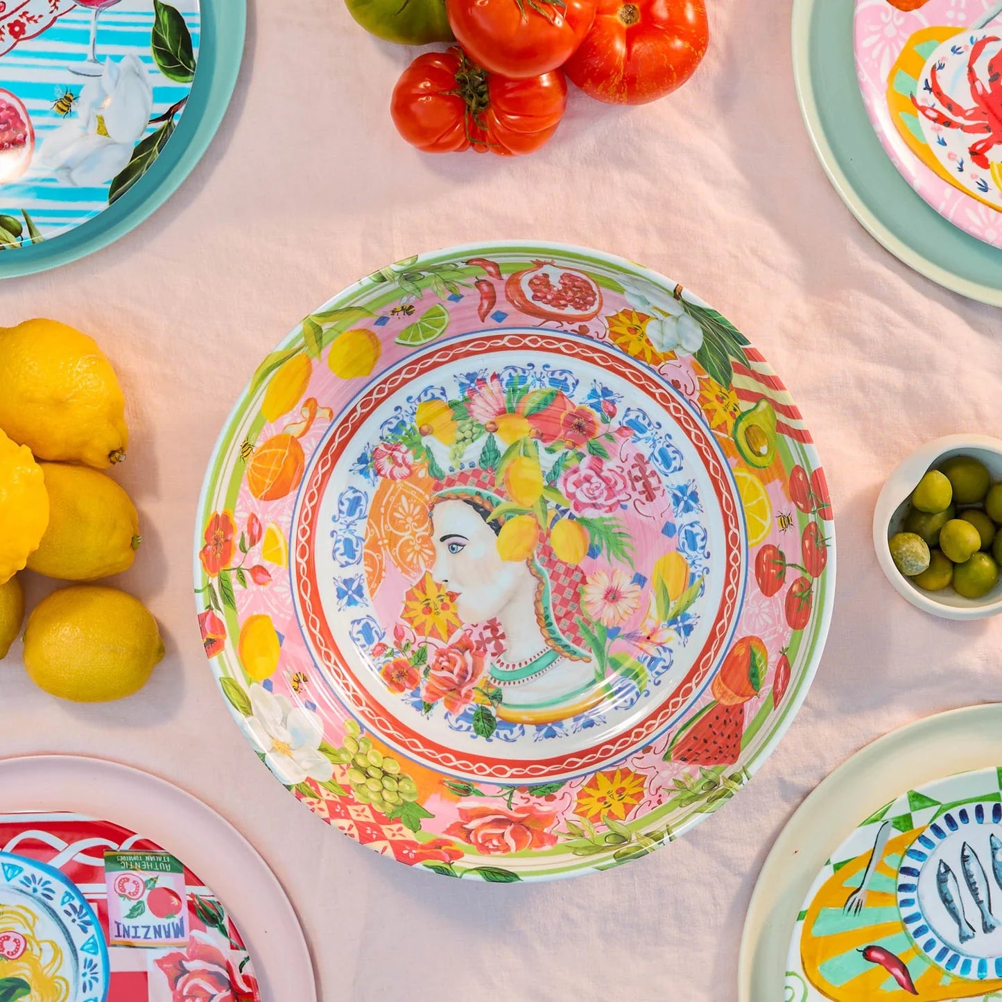 Colourful plates with fruit and floral designs, lemons, tomatoes, and olives on a pink tablecloth.