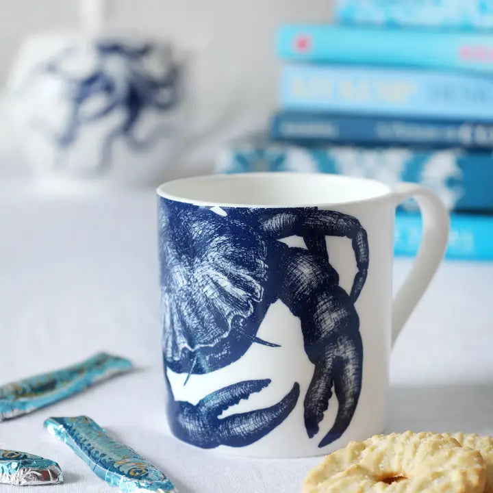Bone china mug with a blue crab illustration, on a white surface with cookies and wrapped chocolates beside it.