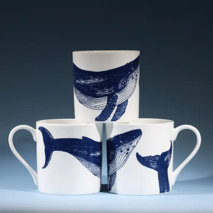 Set of three white ceramic mugs with blue whale art, stacked pyramid-style on dark background.
