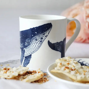 White ceramic mug with blue whale design, displayed on table beside crackers and small plate.