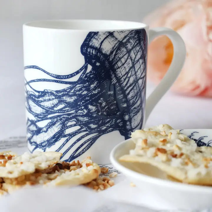 White ceramic mug with blue jellyfish design beside crackers and floral background.