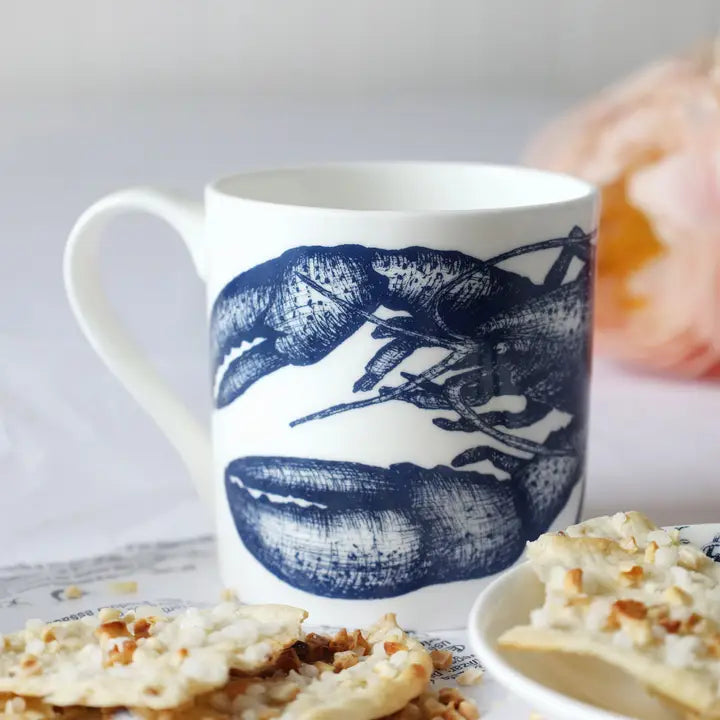 White ceramic mug with blue lobster design on table with crackers and blurred floral background.