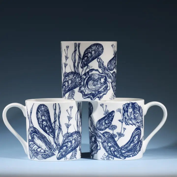 Set of three white ceramic mugs with blue mussel and oyster illustrations, stacked in a pyramid shape.