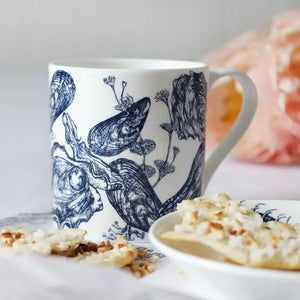 White ceramic mug with blue mussel and oyster illustrations beside a plate of crackers with toppings.