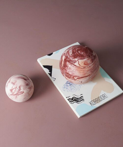 Two spherical objects with a marbled pink and white pattern. One sphere rests on a book titled 'KORRIDOR' with an abstract multicolored cover; the other sphere sits on a mauve surface to the left of the book.