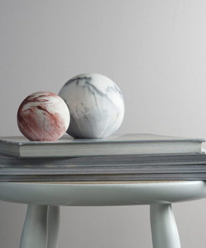 A small red and white marbled sphere and a larger grey and white marbled sphere sit atop a stack of books, which rests on a light-colored round stool with four legs. The background is a plain, light-colored wall.