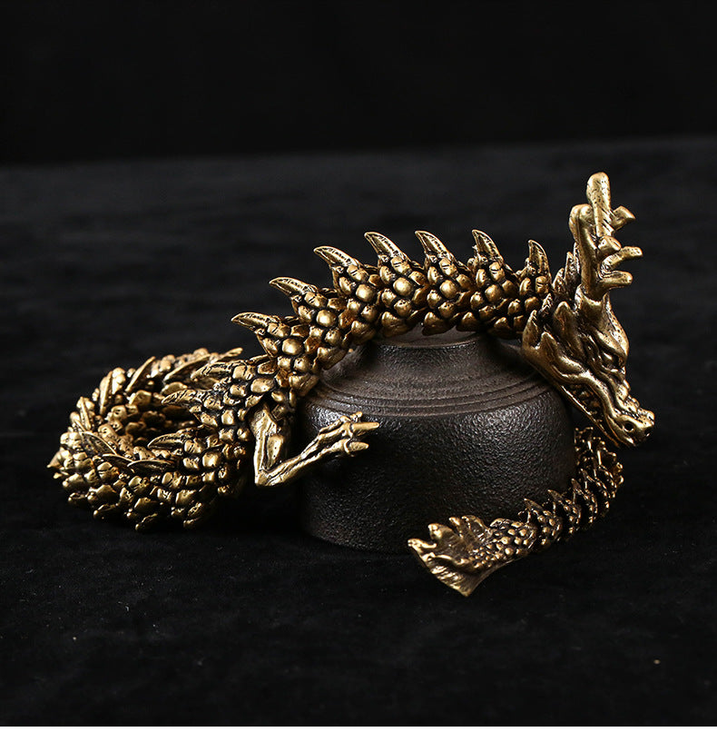 Golden dragon sculpture with a coiled body, intricate scales, sharp claws, and prominent horns. The dragon rests on a black cylindrical pedestal against a black background.
