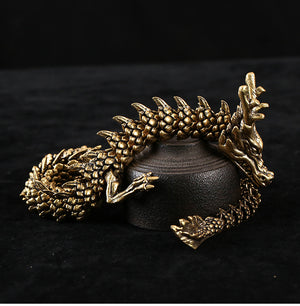 Golden dragon sculpture with a coiled body, intricate scales, sharp claws, and prominent horns. The dragon rests on a black cylindrical pedestal against a black background.