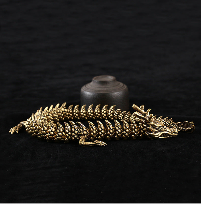 Golden dragon sculpture with intricate scales and a fierce expression, coiled on a black surface. A small dark brown cylindrical object with a lid sits behind it, set against a black background.