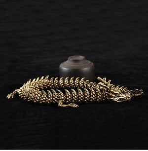 Golden dragon sculpture with intricate scales and a fierce expression, coiled on a black surface. A small dark brown cylindrical object with a lid sits behind it, set against a black background.