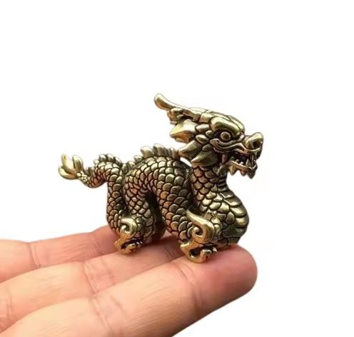 Image of small brass dragon figurine with intricate scale detailing and a fierce expression, shown balanced on a person's fingertips against a white background.