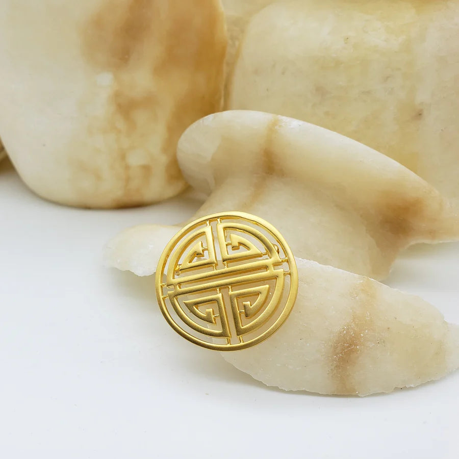 Image of Shou symbol brooch, round gold cut out symbol on white and stone background.