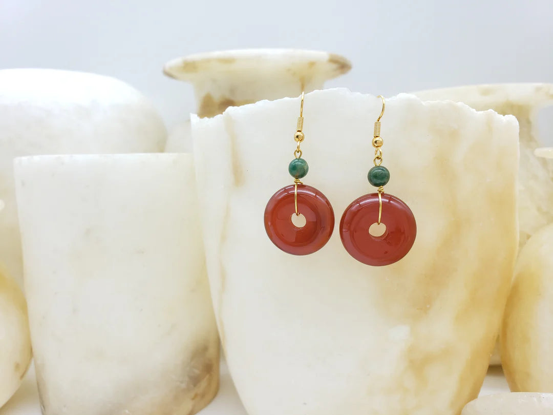 Image of gold pendant earrings with green bead and red bi disc hanging on white crystal.