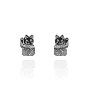 Image of a pair of polished silver stud earrings shaped like lucky cats (Maneki-neko), each with a raised paw and engraved details on the body and face. The earrings are displayed against a white background with a subtle reflection beneath them.