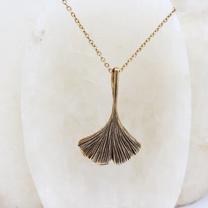 Image of ginko leaf shaped pendant in antique gold on gold chain, hanging on white crystal.