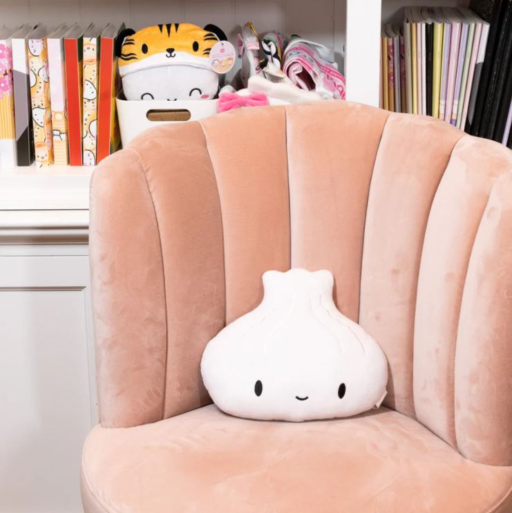 Image of a cute plush toy shaped like a steamed dumpling with a smiling face placed on a soft, peach-colored velvet chair. In the background, there's a white shelf with colorful books, plush toys, and accessories.