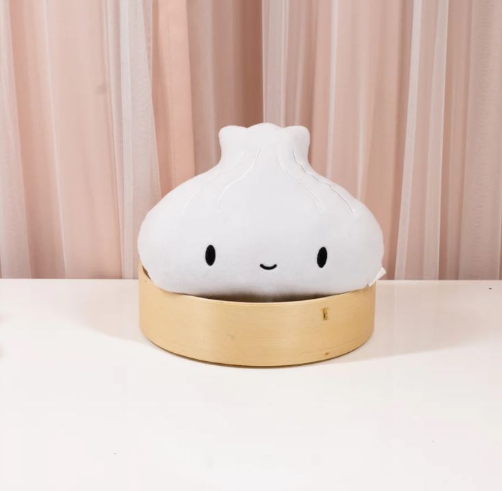 Image of a cute plush toy shaped like a steamed dumpling with a smiling face, sitting inside a round bamboo steamer basket. The background features soft pink and white vertical curtains, and the plush is placed on a white surface.