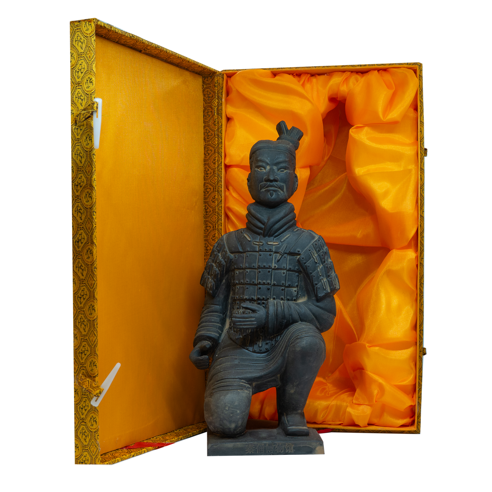 A photo of a kneeling Terracotta Warrior figurine in dark stone, displayed inside an ornate yellow and gold presentation box lined with bright orange satin fabric. The warrior is wearing traditional armour with a topknot hairstyle and has a serious facial expression.