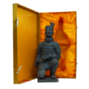 A photo of a kneeling Terracotta Warrior figurine in dark stone, displayed inside an ornate yellow and gold presentation box lined with bright orange satin fabric. The warrior is wearing traditional armour with a topknot hairstyle and has a serious facial expression.