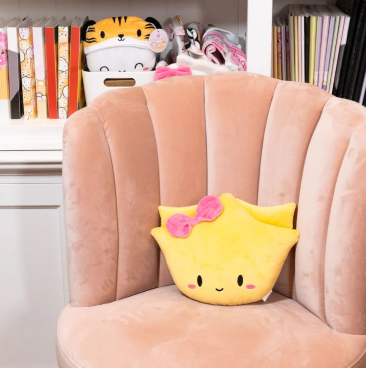 Image of a cute yellow plush toy shaped like a wonton with a smiling face, pink cheeks, and a pink bow on top, resting on a soft peach-colored velvet chair. In the background, a white shelf displays colorful books, a tiger-themed plush container, and various accessories.