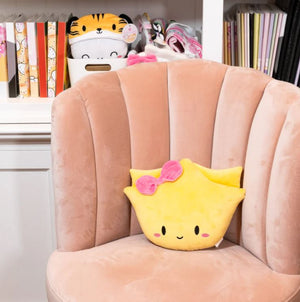 Image of a cute yellow plush toy shaped like a wonton with a smiling face, pink cheeks, and a pink bow on top, resting on a soft peach-colored velvet chair. In the background, a white shelf displays colorful books, a tiger-themed plush container, and various accessories.
