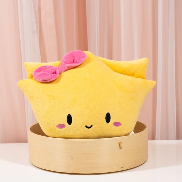 Image of a cute yellow plush toy shaped like a wonton with a smiling face, blushing cheeks, and a pink bow on top. It is sitting inside a round bamboo steamer basket on a white surface, with pink and white curtains in the background.